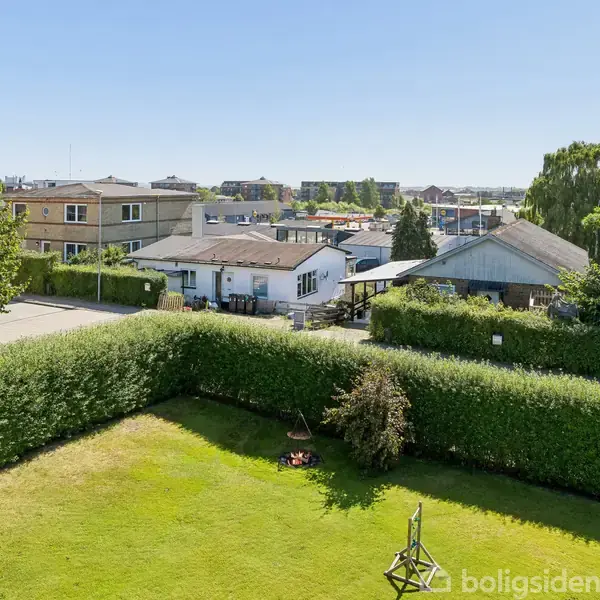 Græsplæne omringet af hæk, stille med en lille busk i midten. I baggrunden ses huse og træer under en klar blå himmel.