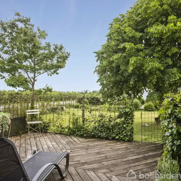 En liggestol står på en træterrasse. Bagved ses en grøn have med store træer, en græsplæne og et hegn. I baggrunden anes en blå himmel.