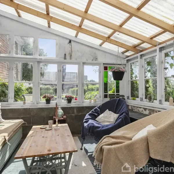 Lys vinterhave med træbænk og bløde puder sidder under et gennemsigtigt tag. Et træbord står i midten, omkranset af vinduer med planter og en statue langs væggen.