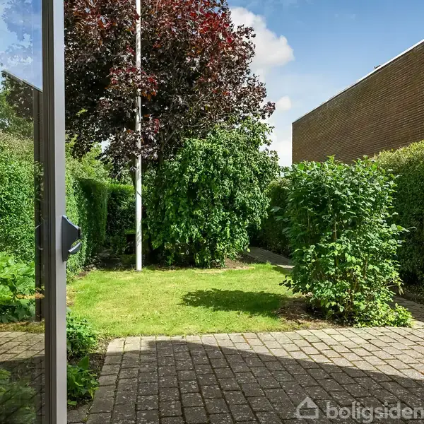 Haveobjektet er en grøn have, hvor buske og et højt træ danner en frodig ramme; en flisebelagt terrasse og murstensbygning ses i baggrunden under en klar himmel.