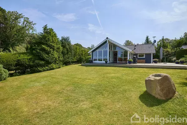 Et moderne hus med store vinduer ligger i en velplejet have med græsplæne og buske. En stor sten er placeret i forgrunden under en klar himmel.