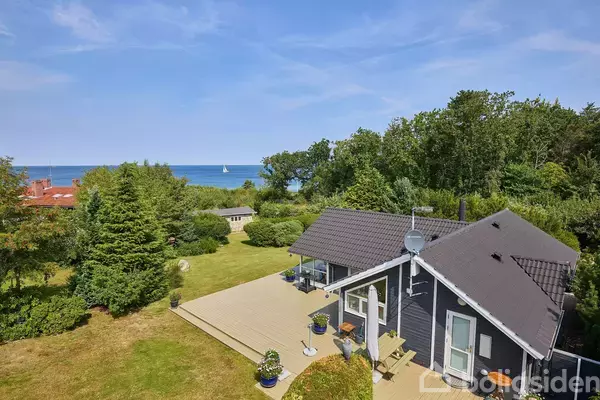 Et mørkt træhus med terrasse står i en frodig have med udsigt til havet i baggrunden, hvor en sejlbåd kan ses. Skov omkranser ejendommen.