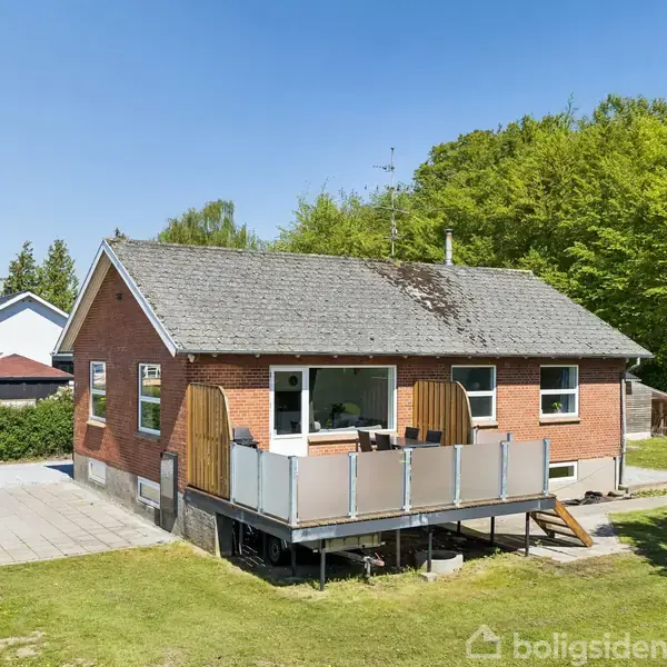 Rødt murstenshus står roligt med en hævet terrasse foran. Sollys belyser huset, omgivet af en grøn have og skovbevoksning i baggrunden, under en klar blå himmel.