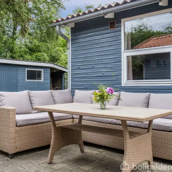 Fletsofa står stille på en terrasse med et bord foran sig og en blomsterdekoration ovenpå. Baggrunden viser et blåt hus med vinduer og en lille skur i en have.