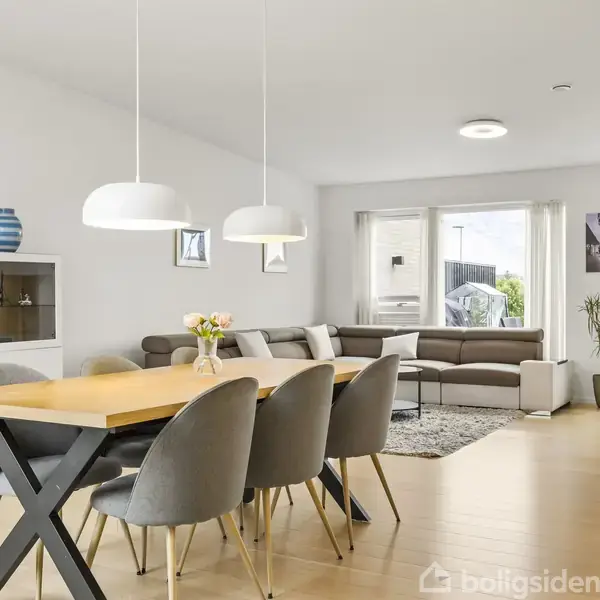 Spisebord med seks polstrede stole står i en lys spisestue. Bagved ses en sofagruppe og et stort vindue med udsigt til have. Moderne indretning med hvide vægge.