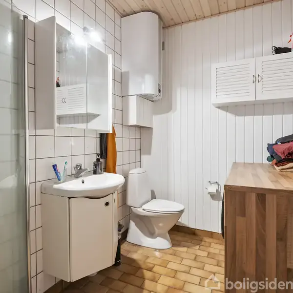 Badeværelse med hvid flisevæg og træloft. Der er et toilet, en håndvask med spejl, et brusehjørne, og en træbordplade med skabe. Håndklæder ligger stablet på bordet.