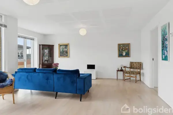 En stue med en blå sofa står centreret i et lyst rum. Til højre ses et mønstret stol og et sidebord. Malerier dekorerer væggene.