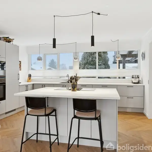 Moderne køkken med en central hvid ø og to sorte barstole, placeret i et lyst rum med store vinduer og hvid vægdekoration.