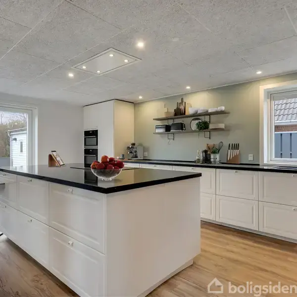 Et moderne køkken med en stor køkkenø fyldt med frugter i en skål. Rummet har lyse trægulve, hvide skabe og indbyggede ovne, med udsigt til en terrasse gennem glasdøre.