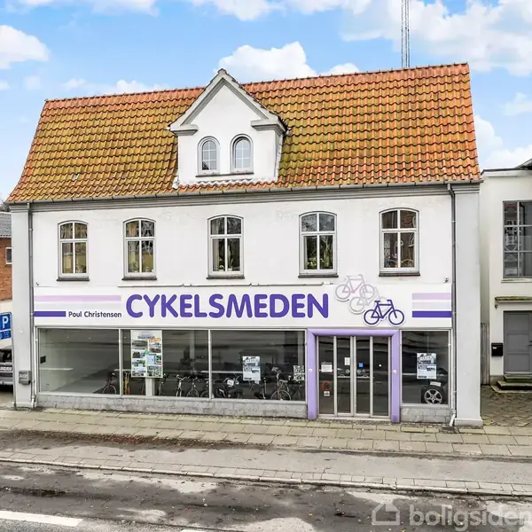 To-etagers bygning med butiksskilte "CYKELSMEDEN" og "Poul Christensen" på facaden. Store vinduer viser cykler indenfor. Bygningen ligger ved en gade med fortov og nabobygninger på begge sider.