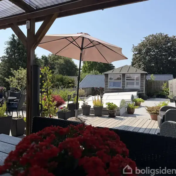 En terrasse med bord og stole, hvor blomster og parasoller skaber en hyggelig atmosfære omgivet af planter og hegn. En mindre bygning ses i baggrunden, omkranset af træer.