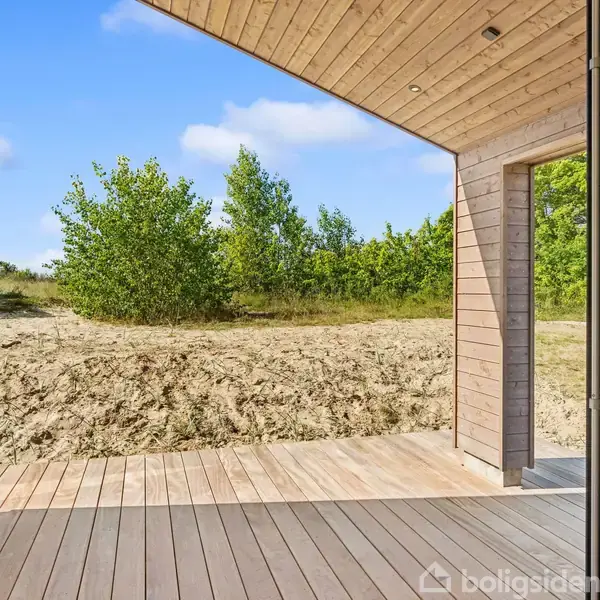 Træterrasse med åbne glasdøre, fører ud til en sandet have med grønne buske i baggrunden under en blå himmel.