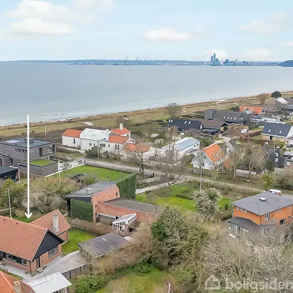 Et hus med rødt tag ses fra luften, placeret i et boligområde nær en kyst. I baggrunden ligger havet og en by ved horisonten.