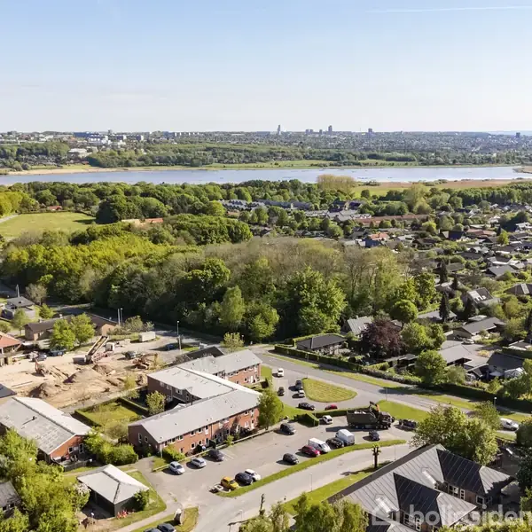 Byområde set fra oven, med huse omkranset af træer. I baggrunden ses en stor sø og en by i horisonten. Klar himmel danner en rolig atmosfære.