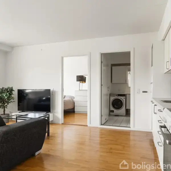 Fladskærms-TV står på glasbordet, sofa foran, i en lys stue med trægulv; døråbning viser vaskemaskine i et rum bagved, køkken til højre.