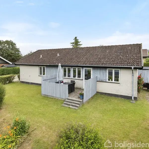 Et enkelt, etplanshus med brun tag og hvid facade står på en grøn græsplæne. Huset har veranda med paraply, omgivet af hegn. Blomsterbede flankerer græsplænen.