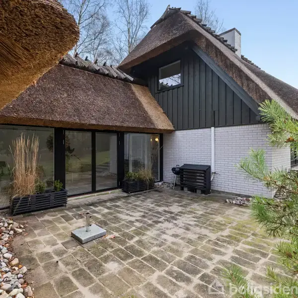 Stråtækt hus med store glaspartier omkranser en flisebelagt gårdhave. Omgivelserne inkluderer dekorative sten og græs. Træer kan ses i baggrunden.