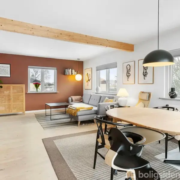 Spisebord og stole står under en sort pendellampe i en stue med træbjælke i loftet. En grå sofa ved vinduer tilbyder udsigt til træer.