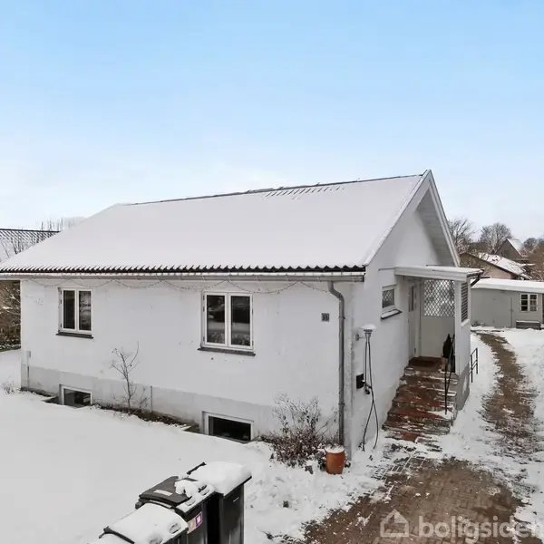 Et hvidt hus med snedækket tag står stille i et vinterlandskab. Huset er omgivet af en snebelagt indkørsel og hæk, og himlen er klar og blå.