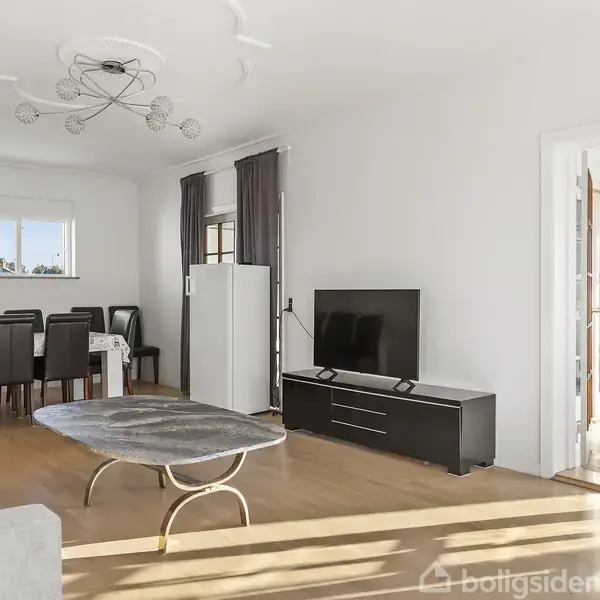 Tv står på et sort mediemøbel i en lys stue, hvor et sofabord og en sofa er placeret. Der er også spisebord og stole i baggrunden.