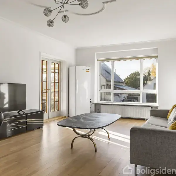 Sofa står stille i lys stue med trægulv og stort vindue. Et sofabord og fladskærms-tv er placeret foran, omgivet af let møblering og indgang med glasdøre.