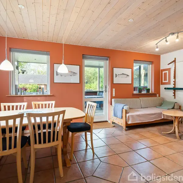 En spisebord med seks stole står under loftslamper i en stue med orange vægge og flisegulv. Der er en sofa og udsigt til en have gennem store vinduer.