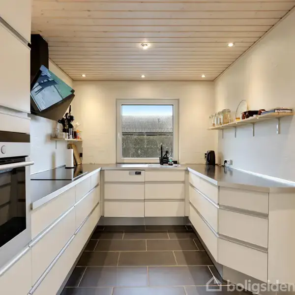 Et moderne køkken med hvide elementer og indbygget ovn, der står i et U-formet layout. Vinduet for enden giver naturligt lys. Loftet er i træ og gulvet i mørke fliser.