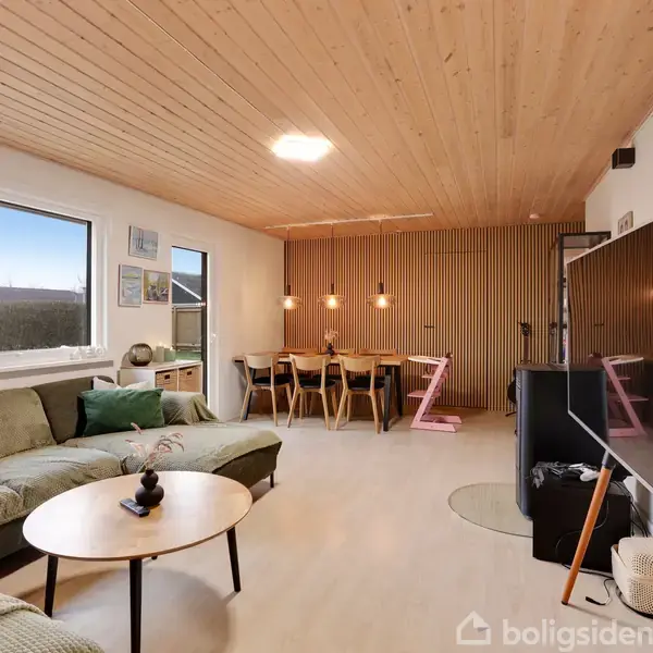 En rummelig stue med en grøn sofa, der står foran et stort vindue. Et rundt sofabord står i forgrunden. I baggrunden ses et spisebord med stole og overhængende lamper.