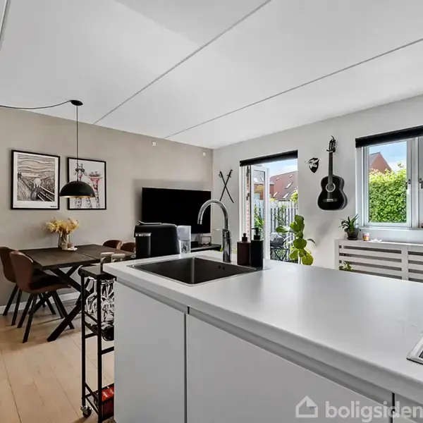 Et moderne køkken-alrum med en hvid køkkenø, spisebord med brune stole, og sorte pendler. En guitar hænger på væggen, og store vinduer giver udsigt til en grøn have.
