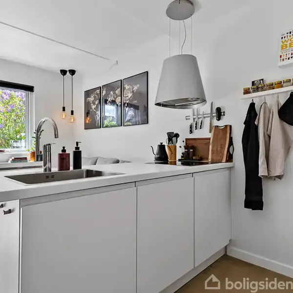 Et moderne køkken med hvidt køkkenbord med indbygget vask. Over bordet hænger en lampe. I baggrunden er et vindue med udsigt til have, og der hænger billeder på væggen.