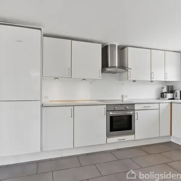 Et moderne køkken med hvide skabe og stålgreb, indbygget ovn og emhætte. Der er en kaffemaskine ved vinduet, der lukker naturligt lys ind. Flisegulv og minimalistisk indretning.