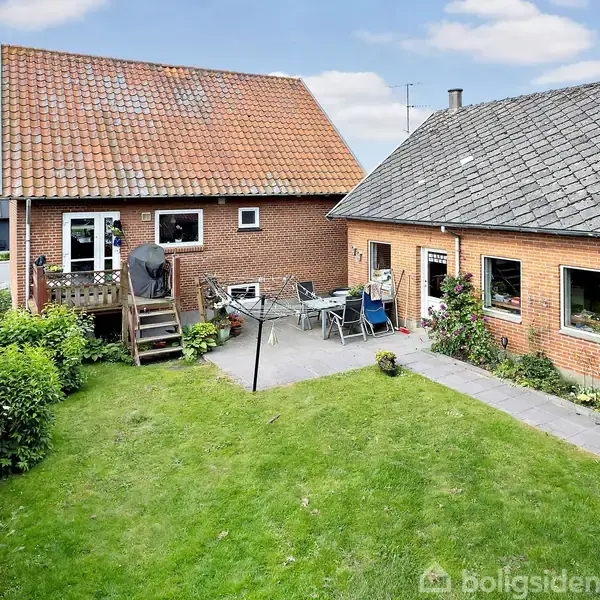 To murstensbygninger står ved siden af hinanden. En terrasse med havemøbler og en grill er omgivet af en grøn græsplæne og buske i en have.