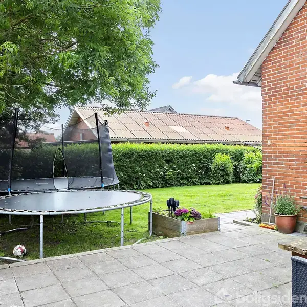 Trampolin med sikkerhedsnet står stille i en græsplæne, omgivet af grønne hække. Nærved står en udendørs ovn på en flisebelagt terrasse ved siden af et rødt murstenshus.