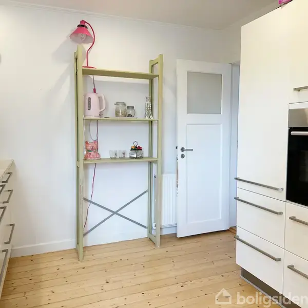 Reol står i hjørnet af køkkenet med en lyserød lampe på toppen og indeholder en rosa kedel, glas og små genstande. Omgivet af hvide vægge og moderne køkkenskabe.