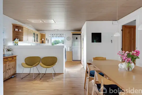 To gule stole står ved en hvid bar i et lyst køkkenområde. Træbord med blomsterbuket og stole ved siden af. Køkkenet har træskabe og hænger i en indbydende, åben rumindretning.