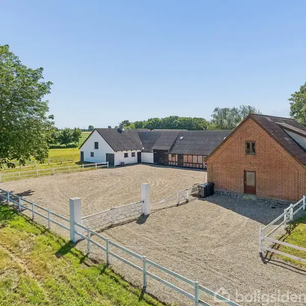 En gård består af en hvid bygning og en rød murstensbygning. Bygningerne omkranser en stor, indhegnet grusplads. Græsmarker og træer omgiver gården i det solrige landskab.