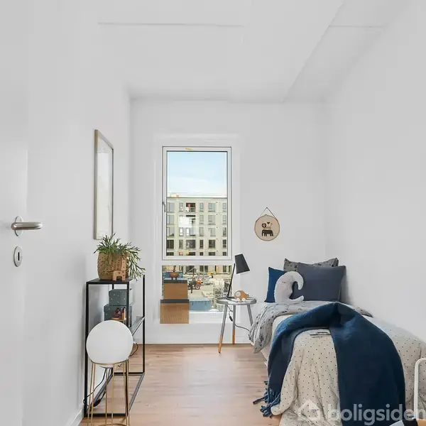 Et lille soveværelse med en enkeltseng dækket med puder og tæppe. Et lille bord med lampe står ved siden af sengen. Et vindue viser moderne bygninger udenfor. En plante står på en reol.