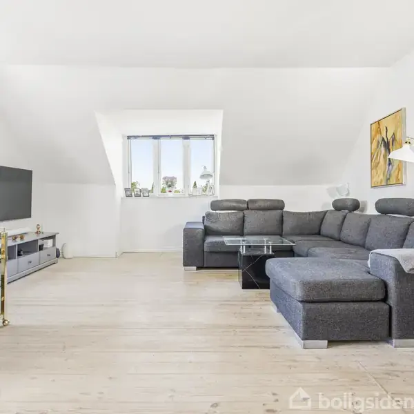 En grå hjørnesofa er placeret foran et sofabord i glas i en lys stue med trægulv. På væggen hænger malerier, og en TV står ved siden af.