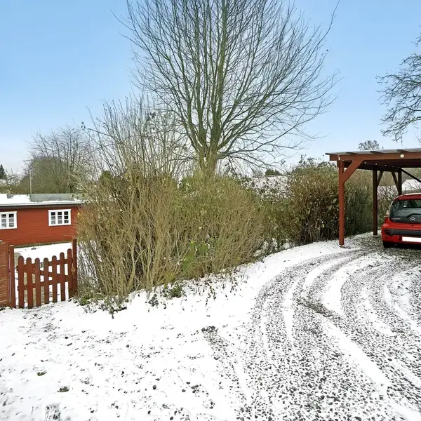 En rød bil holder parkeret under en carport, omgivet af sne og grusvej. I baggrunden ses en rød bygning med et hvidt stakit og buske omkring. Træer står uden blade.
