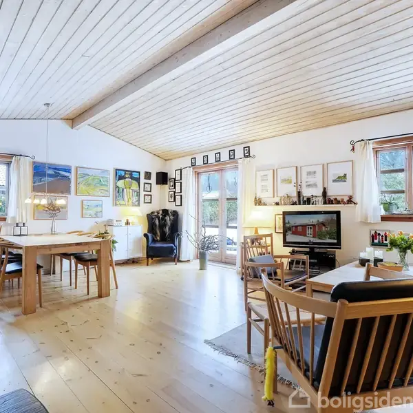 Et lyst, rummeligt stueområde med træmøbler og spisebord til venstre. Store vinduer og glasdøre giver naturligt lys. Der er kunst på væggene, og et TV står til højre.