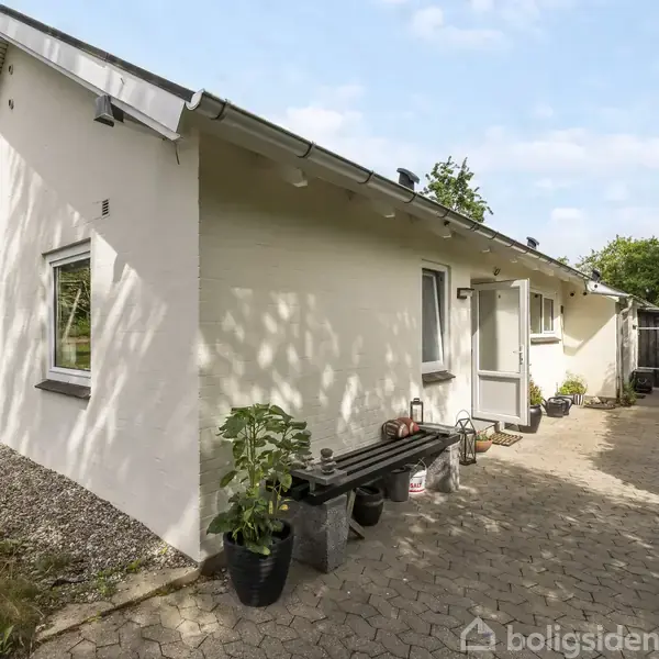 Hvidt murstenshus, set fra siden, med to vinduer og en dør. Foran står en bænk og planter i potter. Omgivet af flisebelagt gang og grønne buske.