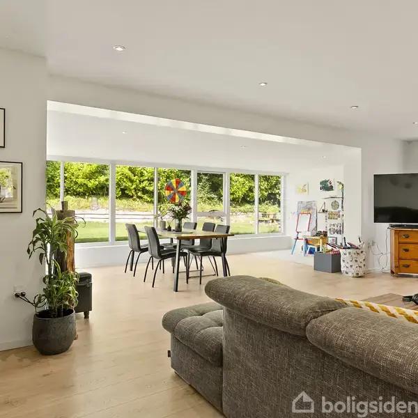 En stue med en grå sofa og et stort tv på en kommode. Spisebord og stole ved store vinduer med udsigt til haven. Planter og kunstværker pynter rummet.