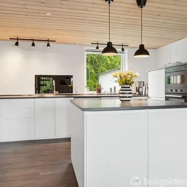 Moderne køkken med hvide skabe og sorte bordplader. Blomster står i en vase på køkkenø, mens indbyggede ovne er til højre. Træloft og store vinduer tilføjer lys og rumsfornemmelse.