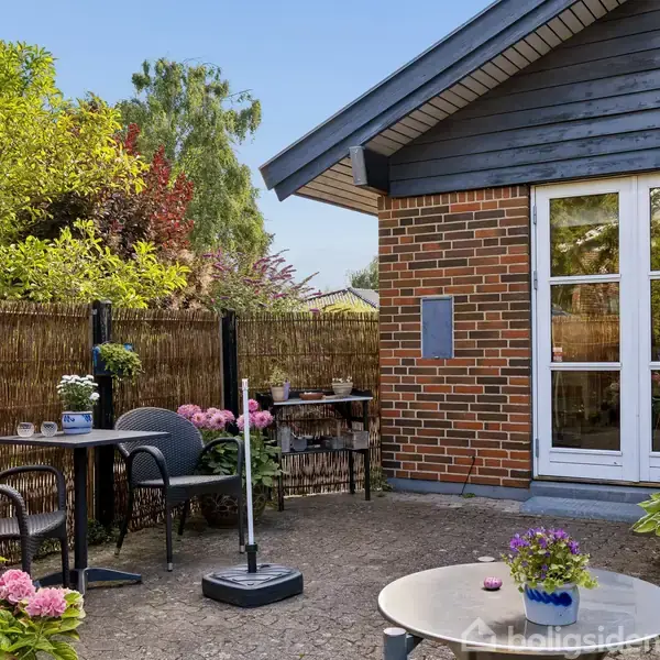 En stenbelagt terrasse har caféborde og stole omgivet af blomstrende hortensiaer, ved siden af et murstenshus med store glasdøre. Baggrunden viser en bambushegn og frodig have.