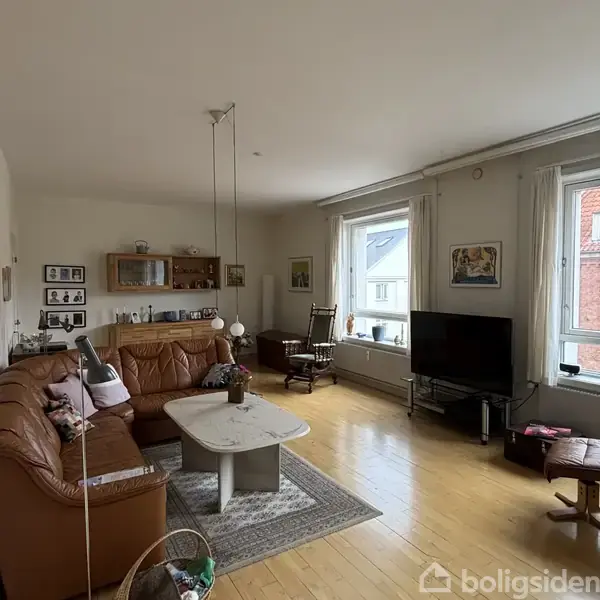 En lys stue med en brun lædersofa og et marmorbord i midten. Et TV står mod væggen på en stander, omgivet af vinduer, reoler og diverse dekorationer.