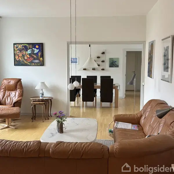 Stue med lædermøbler, hvoraf en lænestol og en sofa står omkring et marmorbord med blomster. I baggrunden ses en spisestue med brune spisebordsstole, vægkunst og hylder.