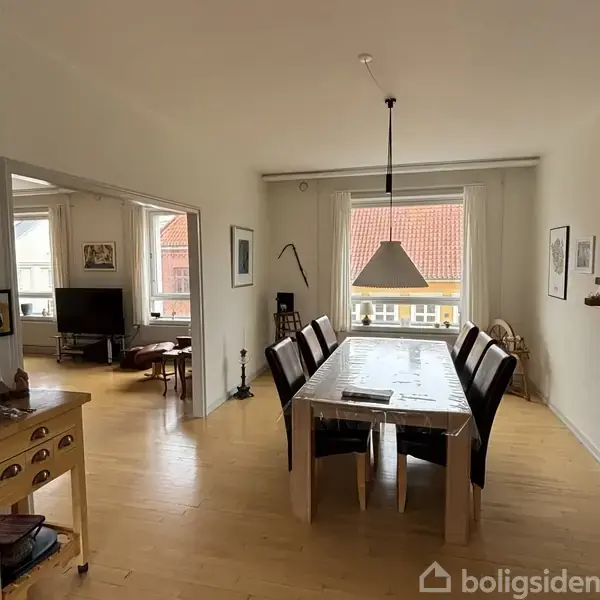 En spisestue med et langt bord dækket med plastikdug omgivet af stole; lampen hænger over bordet. Til venstre ses en åbning til en stue med sofa og TV.