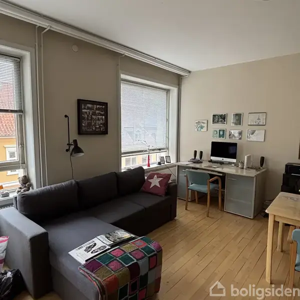 Sofa står ved siden af et stort vindue med persienner. Skrivebordet med computer og højttalere er placeret til højre, omgivet af billeder på væggen. Trægulve dækker rummet.