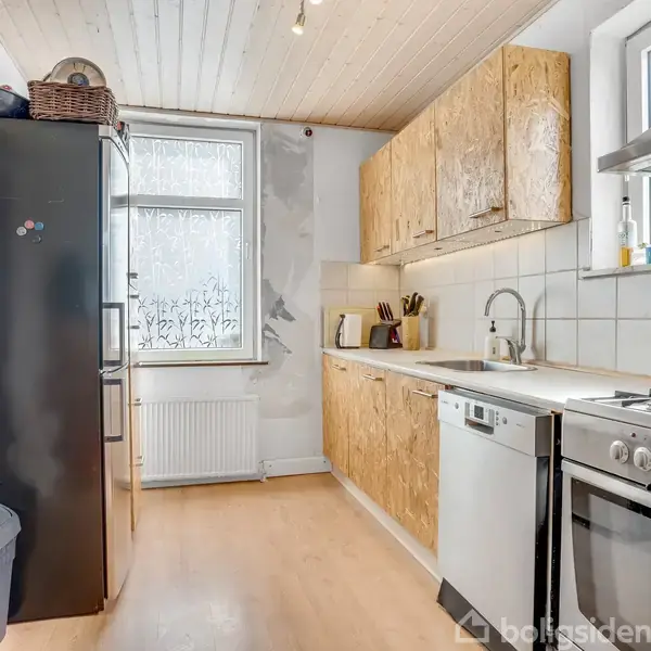 Et køkken med et stort køleskab, opvaskemaskine og komfur langs en væg. Træskabe over og under bordpladen. Lyse trævægge og gulv. Et vindue med frostet glas bagved.