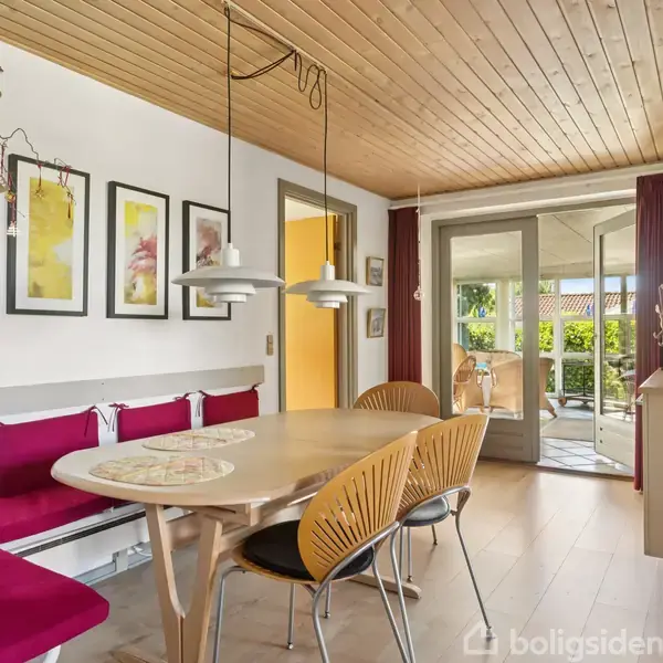 Spisebord med træstole og røde hynder under to hængelamper. Billeder på væggen. Store vinduer og dør åbner mod terrasse med udsigt til have.